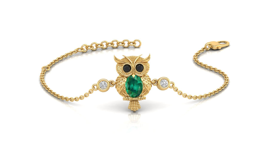 A gold chain bracelet featuring an owl pendant with a green emerald for a beak, black stones for eyes, and two small clear stones on either side, shown on a white background.