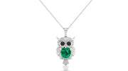 An owl pendant necklace featuring a large green emerald oval cut center stone and small black stones for eyes, set in a white gold metal with tiny clear stones, suspended from a silver chain on a white background.