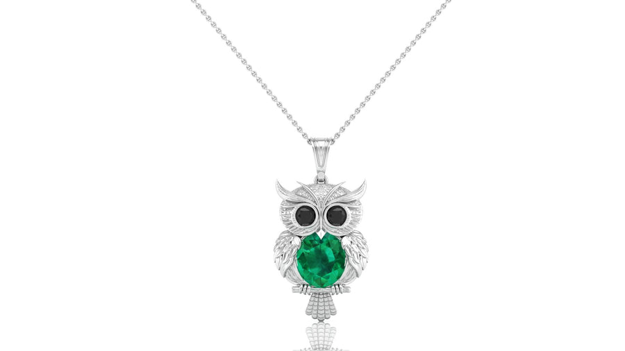 An owl pendant necklace featuring a large green emerald oval cut center stone and small black stones for eyes, set in a white gold metal with tiny clear stones, suspended from a silver chain on a white background.