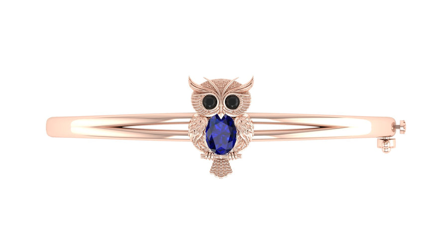 A rose gold bangle bracelet featuring an owl charm with a central Blue sapphire gemstone and small black stone eyes, set against a plain white background.