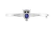 A white gold owl-shaped bangle bracelet featuring a large oval Blue sapphire gemstone in the center, flanked by small black stones for eyes and clear stones for details, set against a plain white background.