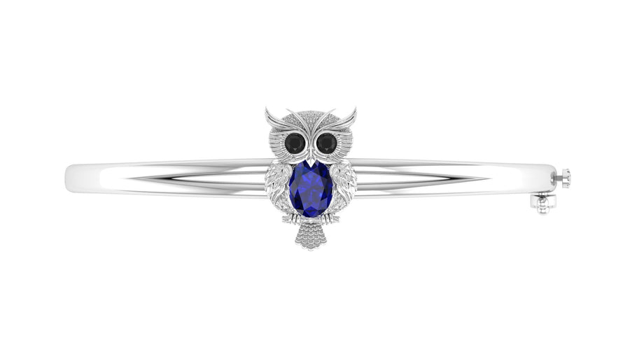 A white gold owl-shaped bangle bracelet featuring a large oval Blue sapphire gemstone in the center, flanked by small black stones for eyes and clear stones for details, set against a plain white background.