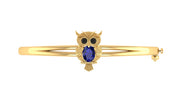 A gold bangle bracelet featuring an owl charm with black eyes and a Blue sapphire body, set against a plain white background.
