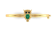 A gold bangle bracelet featuring a central owl charm with a green emerald-cut face and black stone eyes, shown on a plain white background.