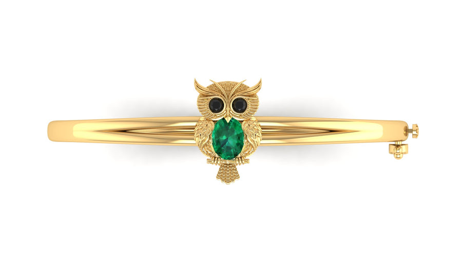 A gold bangle bracelet featuring a central owl charm with a green emerald-cut face and black stone eyes, shown on a plain white background.