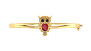 A gold bangle bracelet featuring a central owl charm with black eyes and a red ruby gemstone on the centre, set against a plain white background.