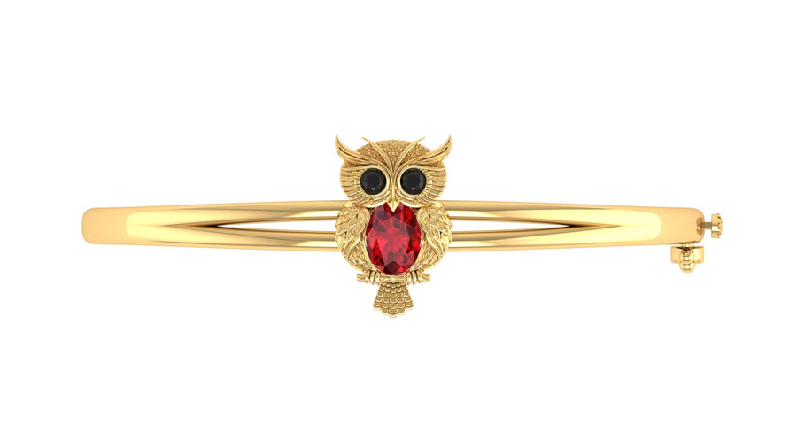 A gold bangle bracelet featuring a central owl charm with black eyes and a red ruby gemstone on the centre, set against a plain white background.