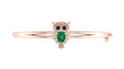 A rose gold bangle bracelet featuring an owl charm with a green emerald-cut stone and black stone eyes, set against a plain white background.