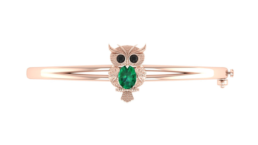 A rose gold bangle bracelet featuring an owl charm with a green emerald-cut stone and black stone eyes, set against a plain white background.